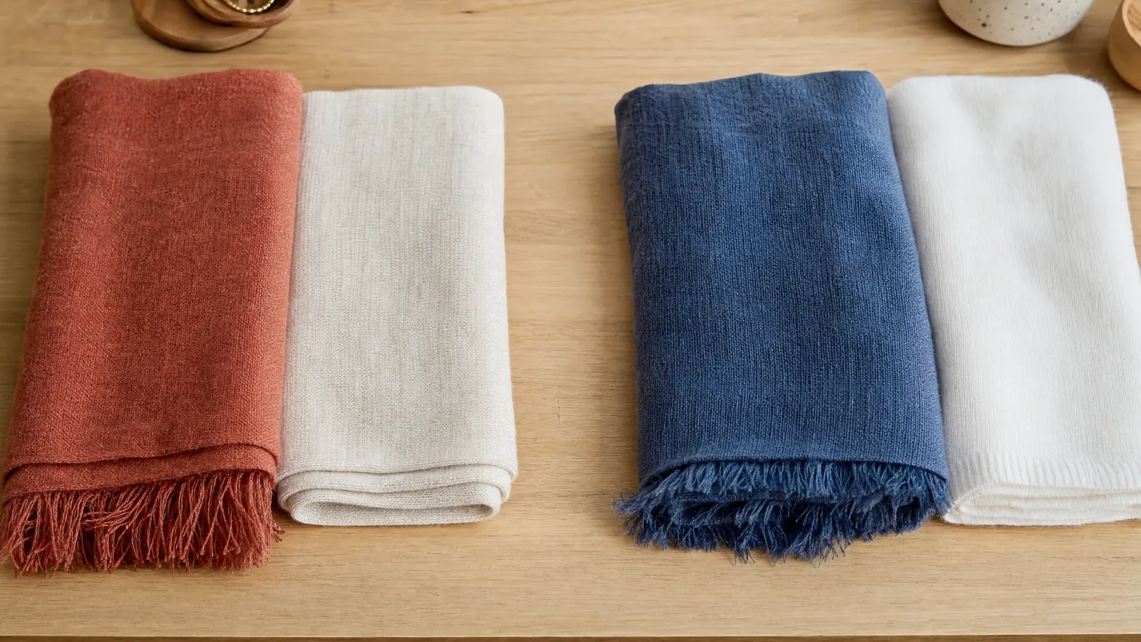Folded terracotta and warm ivory scarves beside smoky blue and white scarves on a neutral wooden surface.