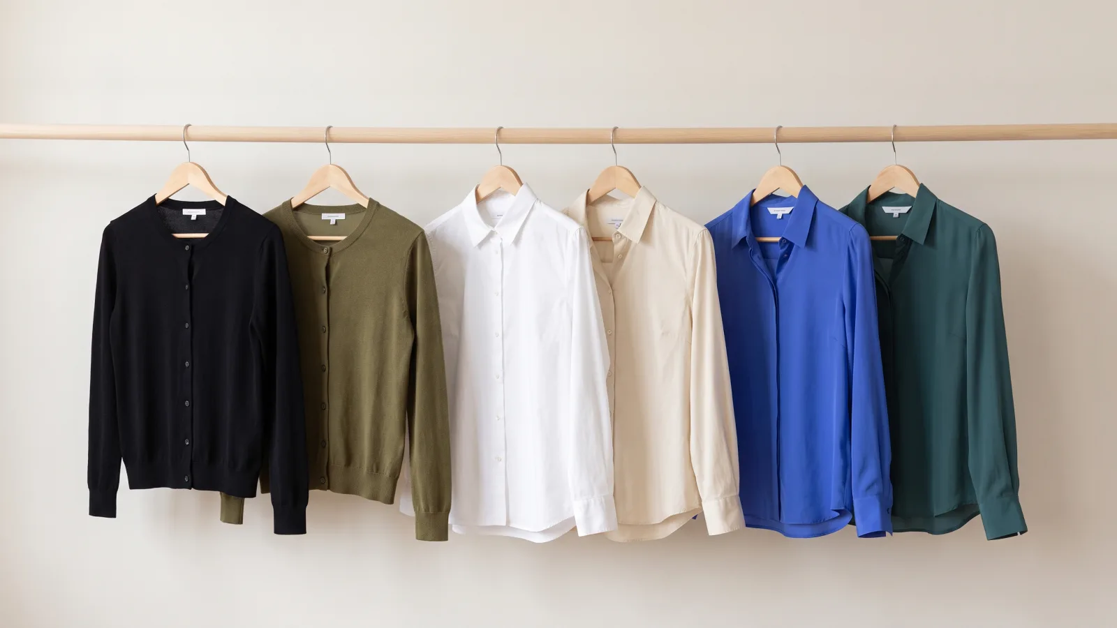 Six tops on a wooden closet rail in three pairs, a black cardigan beside an olive cardigan, a bright white shirt beside a warm ivory shirt, and a cobalt blouse beside a deep teal blouse.