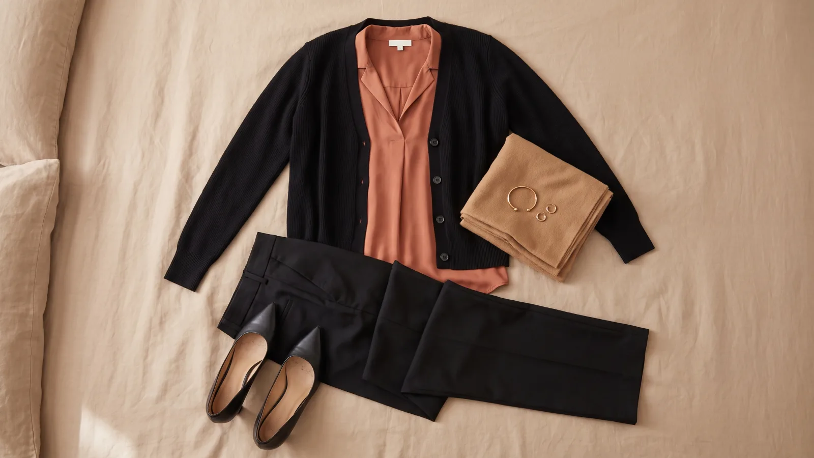 A black cardigan lies open over a terracotta silk blouse on a linen bed with black trousers below it and a camel scarf beside it.